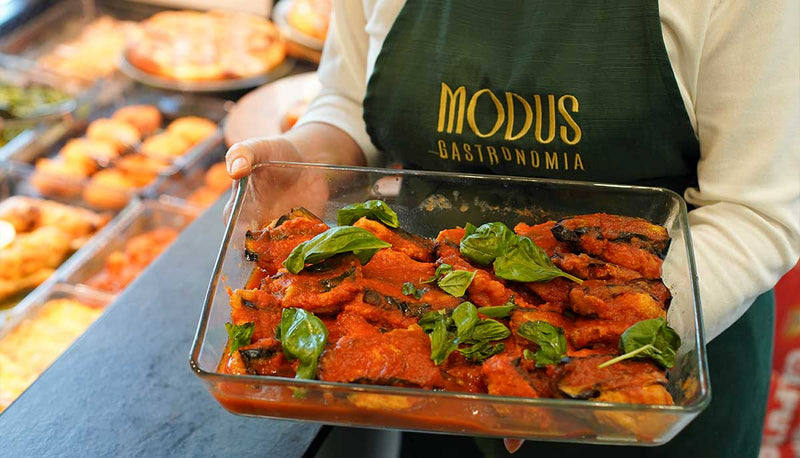 Teglia di melanzane al pomodoro con basilico, tenuta da un addetto con grembiule Modus